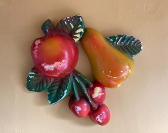 May include: A ceramic wall decor featuring a red apple, an orange pear, and two red cherries with green leaves.