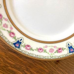 May include: A white porcelain plate with a gold rim and a floral design. The design features pink roses, blue and green leaves, and a gold chain pattern.