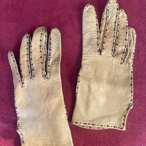 May include: A pair of light tan leather gloves with dark brown stitching along the seams. The gloves have a classic design, with individual fingers and a wrist opening. The leather appears to be soft and supple, suggesting a vintage or high-quality item.