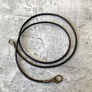 May include: A dark brown leather necklace cord with a clasp and a small loop. The cord is coiled in a circular pattern. The clasp and loop are a brass colour. The necklace is on a grey textured surface.
