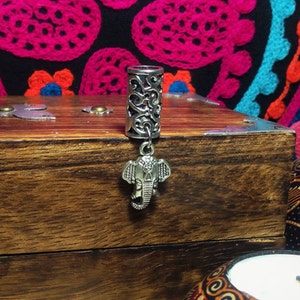May include: Silver hair tie with an elephant charm. The hair tie is on a wooden box with a latch. The box is in front of a colourful fabric with a circular pattern.