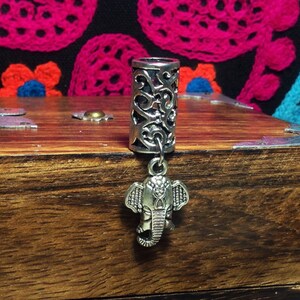 May include: Silver hair tie with an intricate design and an elephant charm. The hair tie is on a wooden surface.