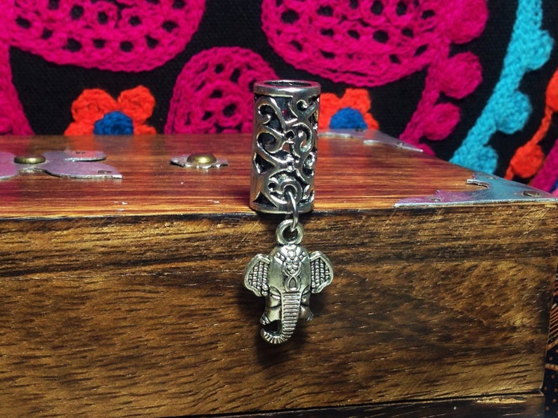 May include: Silver hair tie with an intricate design and an elephant charm. The hair tie is on a wooden surface.