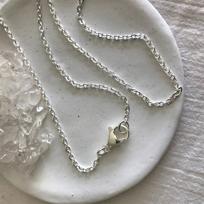 Op de afbeelding: Een zilveren ketting met een karabijn sluiting. De ketting ligt op een wit oppervlak.