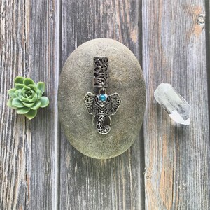 K&ouml;nnte beinhalten: Silberne Haarklammer mit Elefantenmotiv und einem blauen Edelsteinauge. Die Klammer liegt auf einem grauen Stein.