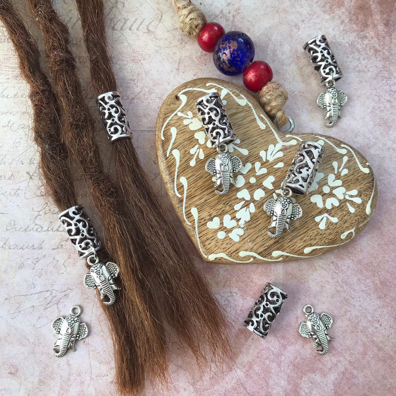 May include: Six silver dreadlock beads with an elephant charm. The beads are on a wooden heart-shaped surface and on brown dreadlocks.