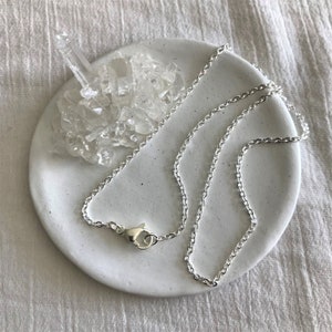 Op de afbeelding: Een zilveren ketting met een karabijn sluiting. De ketting ligt op een wit keramiek schaaltje met heldere kwartskristallen.