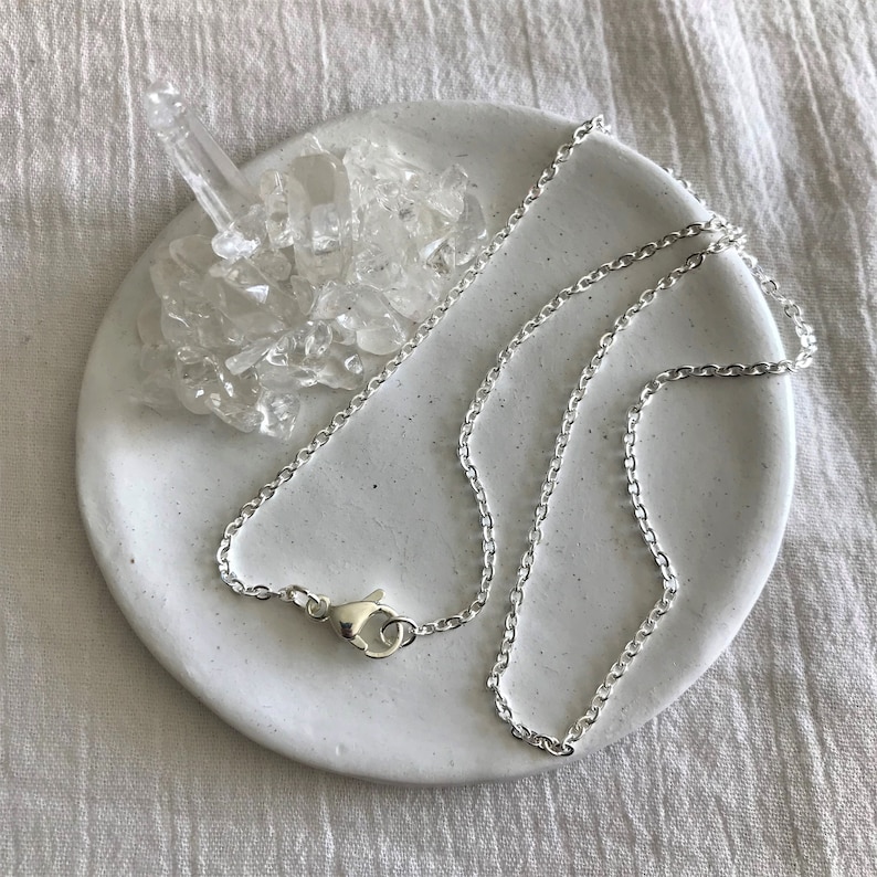 Op de afbeelding: Een zilveren ketting met een karabijn sluiting. De ketting ligt op een wit keramiek schaaltje met heldere kwartskristallen.