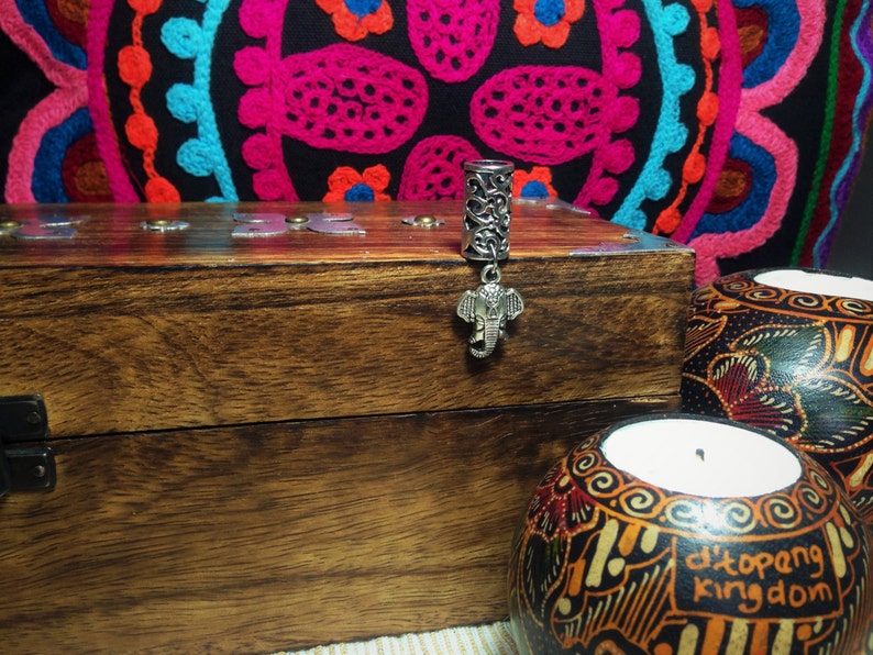 May include: Silver hair tie with an elephant charm. The hair tie is on a wooden box with a latch. The box is in front of a colourful fabric with a circular pattern.