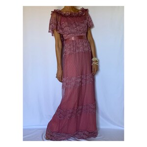 Puede incluir: Un vestido largo de encaje color rosa polvoriento con un escote con volantes y una faja de sat&eacute;n. El vestido tiene una falda de capas y mangas cortas.
