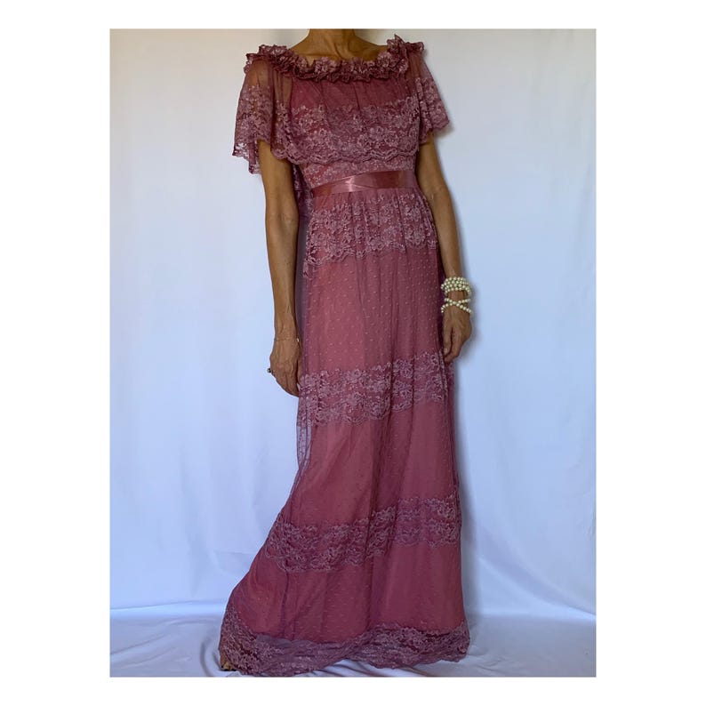 Puede incluir: Un vestido largo de encaje color rosa polvoriento con un escote con volantes y una faja de sat&eacute;n. El vestido tiene una falda de capas y mangas cortas.