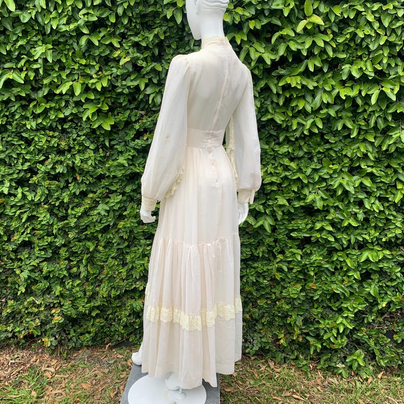Puede incluir: Un vestido maxi largo blanco con cuello alto y mangas largas. El vestido tiene una falda de capas y ribete de encaje. El vestido est&aacute; hecho de una tela ligera y es perfecto para una boda de verano u otra ocasi&oacute;n especial.