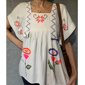 May include: A white blouse featuring colourful cross-stitch floral embroidery in shades of neon pink, orange, blue, and green. The garment has a square neckline, short flutter sleeves, and a brown leather shoulder strap visible on the side.