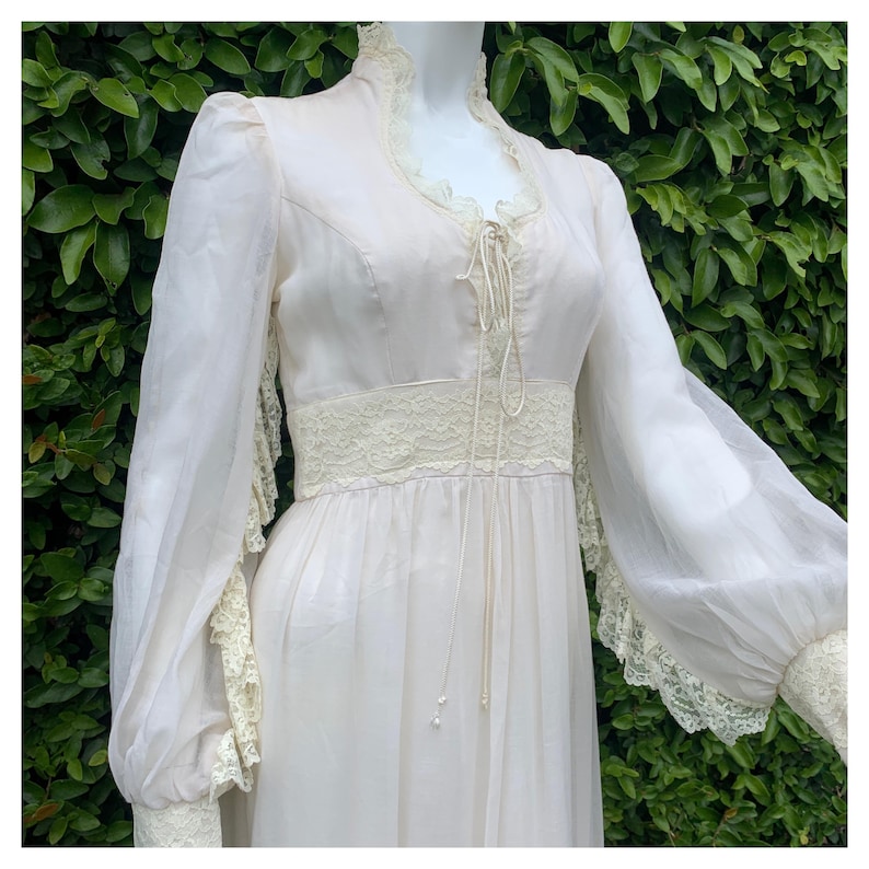 Puede incluir: Un vestido de novia vintage blanco con mangas largas y detalles de encaje. El vestido tiene un borde de encaje alrededor del escote y la cintura, y las mangas est&aacute;n acabadas con pu&ntilde;os de encaje.