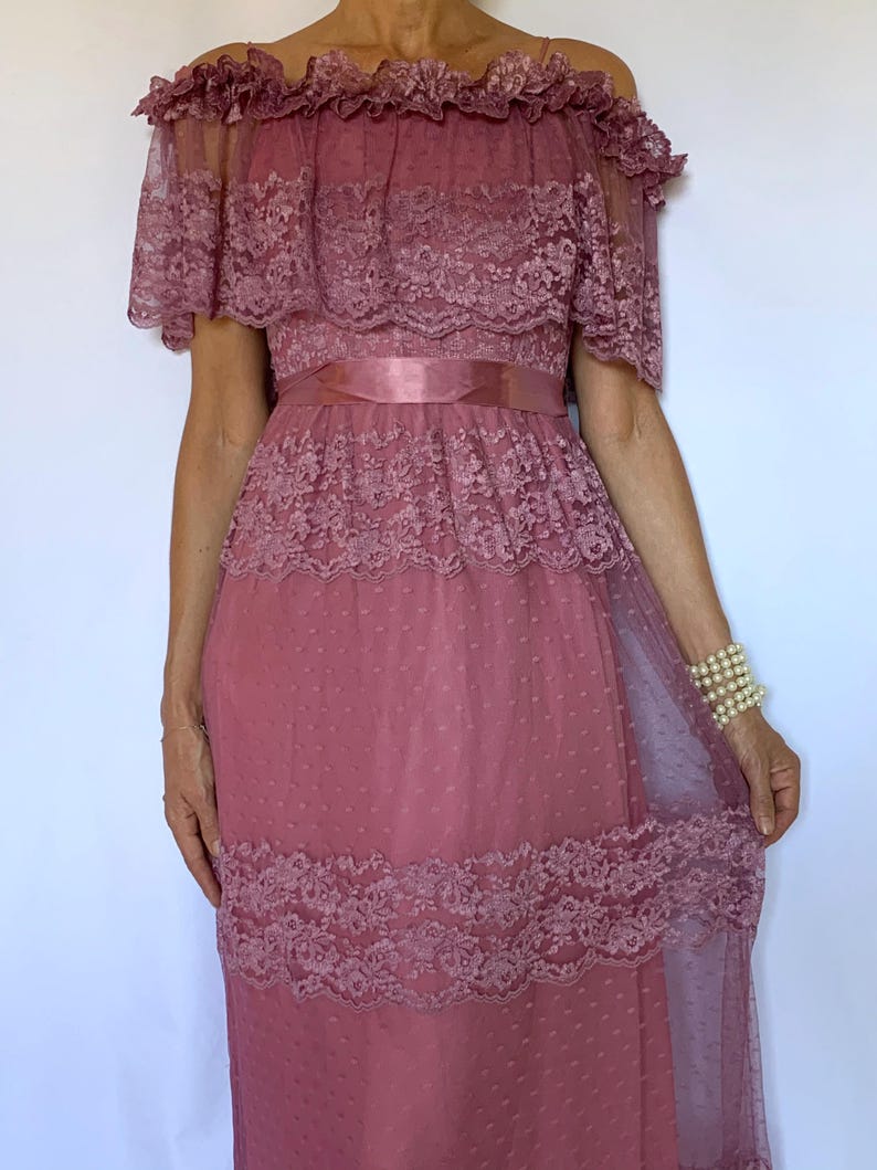 Puede incluir: Un vestido largo de color rosa, de hombros descubiertos, con detalles de encaje y una cinta de raso en la cintura.