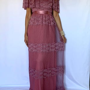Puede incluir: Un vestido largo de encaje color malva con hombros descubiertos y una cinta de raso en la cintura. El vestido tiene una tela transparente con lunares y ribetes de encaje.