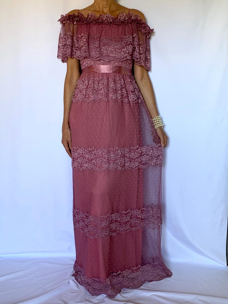 Puede incluir: Un vestido largo de encaje color malva con hombros descubiertos y una cinta de raso en la cintura. El vestido tiene una tela transparente con lunares y ribetes de encaje.
