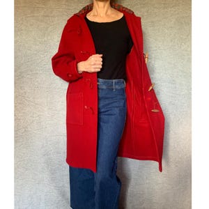 May include: A vibrant red duffel coat featuring wooden toggles and a plaid-lined hood. The coat has two front pockets and is open, revealing a black top and blue jeans. A classic outerwear garment.