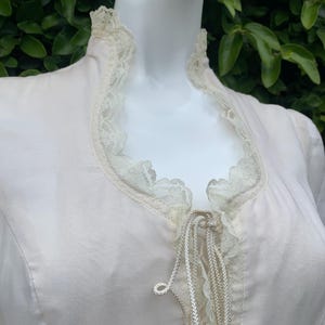 Puede incluir: Una blusa blanca con un ribete de encaje alrededor del escote y un lazo de encaje en la parte delantera. El lazo est&aacute; atado en un lazo.