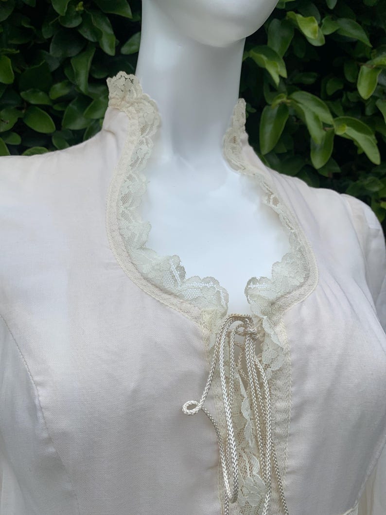 Puede incluir: Una blusa blanca con un ribete de encaje alrededor del escote y un lazo de encaje en la parte delantera. El lazo est&aacute; atado en un lazo.