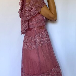 Puede incluir: Un vestido maxi rosa polvoriento con una superposici&oacute;n de encaje y un escote con volantes. El vestido tiene un corpi&ntilde;o ajustado y una falda fluida.