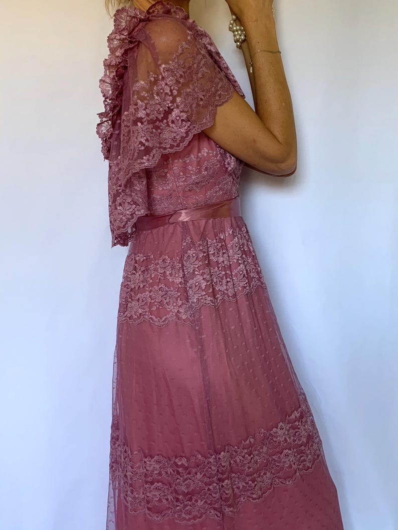 Puede incluir: Un vestido maxi rosa polvoriento con una superposici&oacute;n de encaje y un escote con volantes. El vestido tiene un corpi&ntilde;o ajustado y una falda fluida.