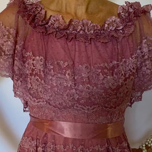 Puede incluir: Un vestido de encaje rosa polvoriento con un escote de volantes y una faja de sat&eacute;n a juego. El vestido tiene mangas cortas y una falda fluida.