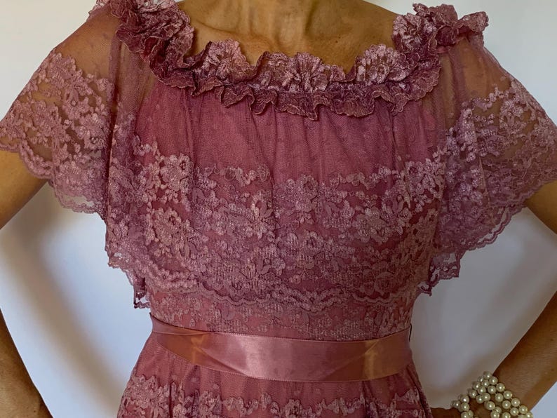 Puede incluir: Un vestido de encaje rosa polvoriento con un escote de volantes y una faja de sat&eacute;n a juego. El vestido tiene mangas cortas y una falda fluida.