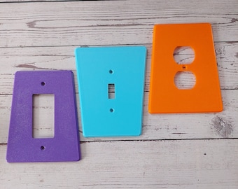 May include: Three plastic wall plate covers in different colors and styles. A purple cover with a single light switch opening, a light blue cover with a single light switch opening, and an orange cover with a double outlet opening.