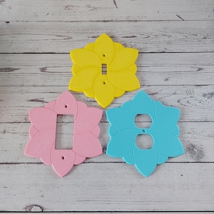 May include: Three decorative light switch and outlet covers in the shape of a flower. The covers are yellow, pink, and blue.