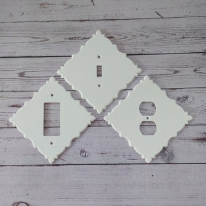 Puede incluir: Tres placas de pared decorativas blancas con diseños intrincados. Las placas son para interruptores de luz y tomas de corriente. Las placas están sobre un fondo de madera clara.