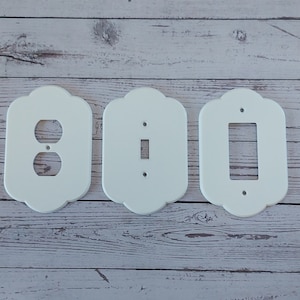 May include: Three white plastic wall plates with decorative edges. The plates are for electrical outlets and light switches. The plates are on a white wood background.