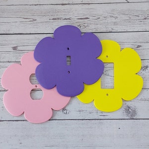 May include: Three flower-shaped light switch covers in pink, purple, and yellow. The covers are made of plastic and have a smooth finish.