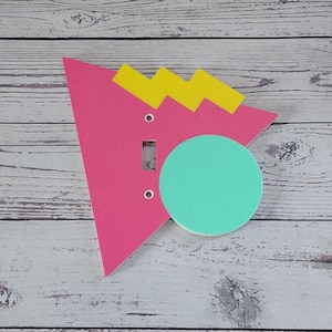May include: A pink, yellow, and turquoise geometric light switch cover with a circle and zig-zag design.