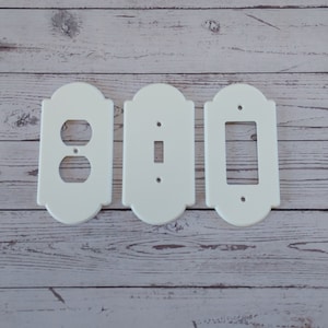 Può includere: Tre placche da parete in plastica bianca con bordi decorativi. Le placche sono per prese elettriche e interruttori della luce. Le placche sono su uno sfondo in legno chiaro.