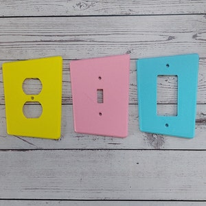 Puede incluir: Tres placas de pared decorativas de estilo retro. Las placas son amarillas, rosas y azules y presentan un solo recorte para interruptor o toma de corriente.