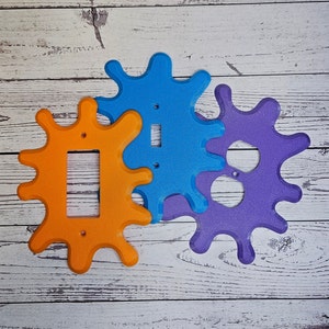 May include: Three colourful light switch covers in the shape of a splatter or blob. The covers are orange, blue, and purple. The orange cover has a single light switch opening, the blue cover has a single light switch opening, and the purple cover has a double light switch opening.