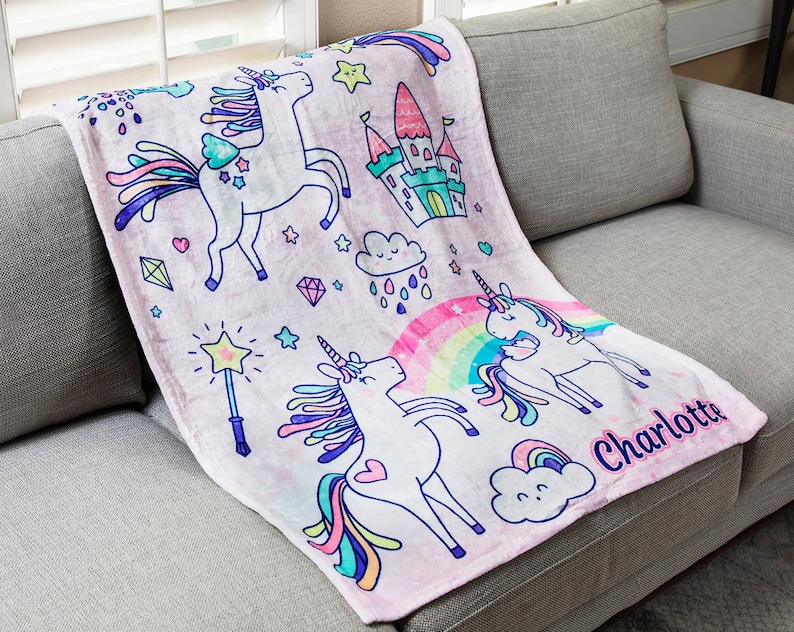 May include: Pink fleece blanket with a unicorn, rainbow, and castle design. The blanket has the name "Charlotte" printed on it.