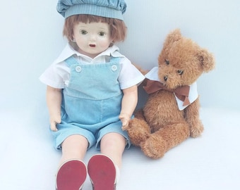 May include: A vintage porcelain doll wearing a blue and white striped overall outfit and a blue and white striped hat. The doll is sitting with a brown teddy bear wearing a white collar with a brown bow.