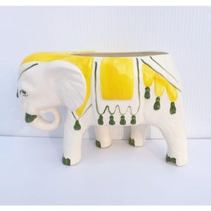 May include: A white ceramic elephant planter with yellow and green accents. The elephant has a yellow saddle and decorative green tassels. The planter is designed to hold plants and adds a whimsical touch to any space.