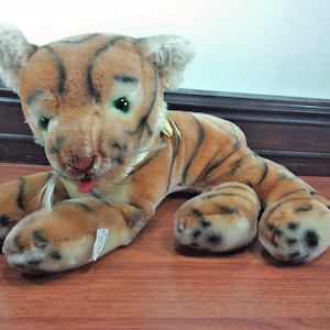 May include: A plush tiger toy with brown and black stripes, green eyes, and a pink tongue sticking out. The toy is lying on a wooden surface.