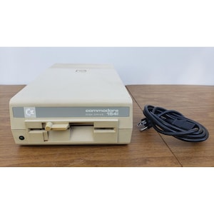 May include: A beige Commodore 1541 disk drive with a rectangular casing, a front-loading slot, a manual latch lever, and a small green power indicator light. The front panel features the text "commodore DISK DRIVE 1541". A black power cord is included.