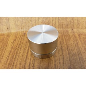 May include: A cylindrical metal knob with a smooth, flat top surface featuring a radial brushed finish and a textured, vertically grooved side band. The object has a metallic silver color and a solid, compact construction with a circular profile.