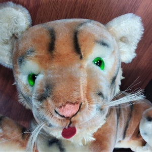 May include: A plush tiger toy with brown and black stripes, white fur, and green eyes. The tiger has a pink nose and a red tongue sticking out.