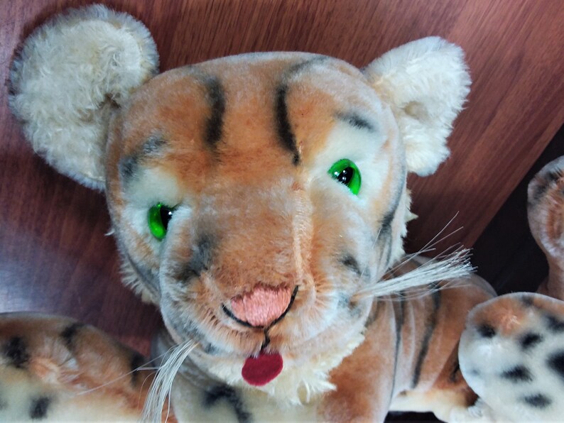 May include: A plush tiger toy with brown and black stripes, white fur, and green eyes. The tiger has a pink nose and a red tongue sticking out.