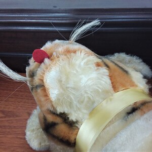 May include: A plush tiger toy with orange and black stripes, white fur, and a red nose. The toy has a yellow ribbon around its neck.