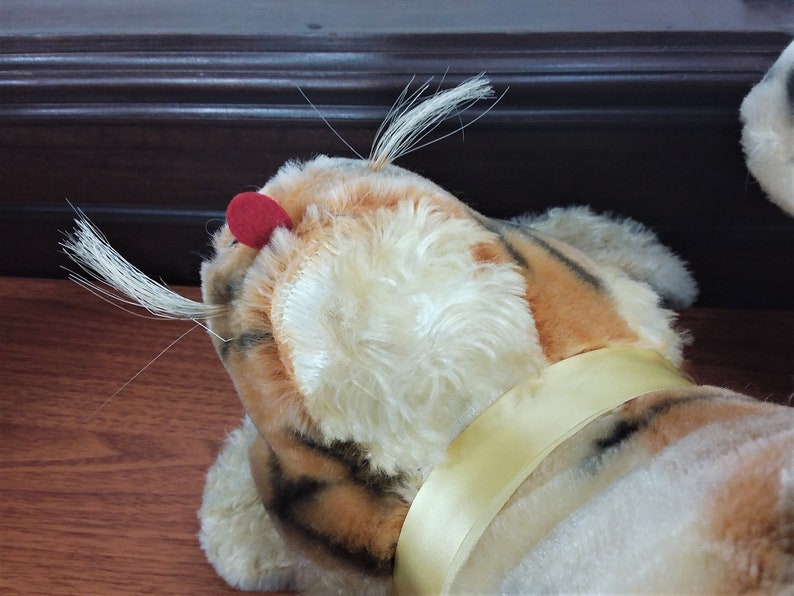 May include: A plush tiger toy with orange and black stripes, white fur, and a red nose. The toy has a yellow ribbon around its neck.