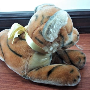 May include: A plush tiger toy with orange and black stripes, lying on its side with a yellow satin ribbon around its neck. The toy has a realistic appearance with a soft, furry texture.