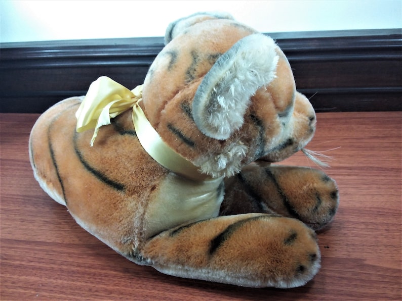 May include: A plush tiger toy with orange and black stripes, lying on its side with a yellow satin ribbon around its neck. The toy has a realistic appearance with a soft, furry texture.