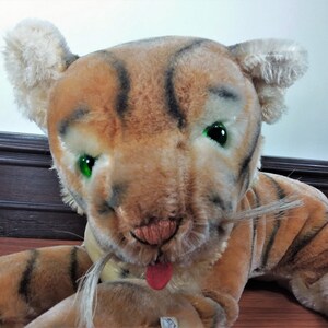 May include: A plush tiger toy with brown and black stripes, green eyes, and a pink tongue sticking out. The toy has a white mustache and is lying on a wooden floor.